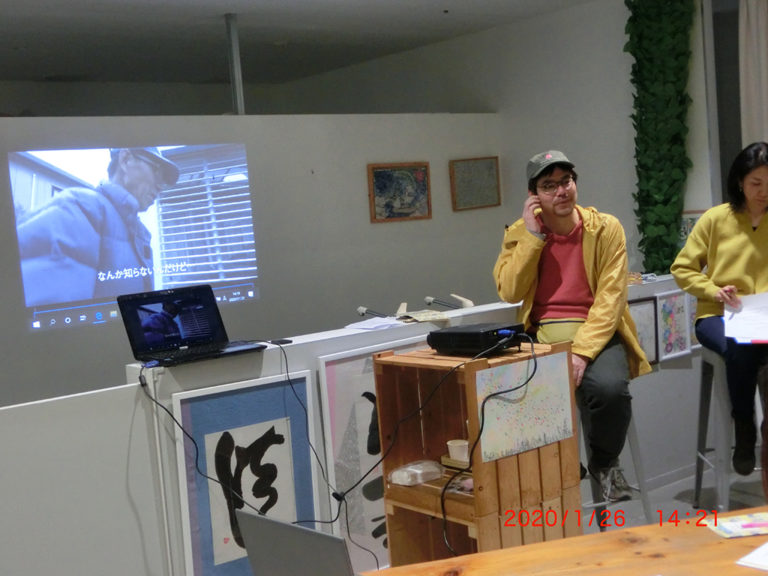 写真です。坪田監督がハイスツールに座りながらお話しています。スライドを背にし、隣には司会進行の吉川さんがいます。